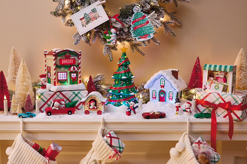 Festive holiday mantel display featuring miniature Christmas village decorations, wrapped gifts with plaid patterns and red ribbon, a decorated Christmas tree, toy cars, and snowy houses. Above the mantel, two holiday card ornaments hang from a flocked tree branch, including a Christmas tree-shaped design with the word “Christmas” and a card illustrated with a festive storefront. Knit stockings filled with gifts hang below, completing the cozy holiday scene.