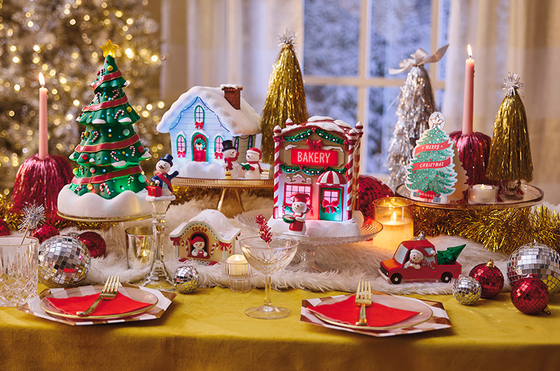 Close-up holiday display featuring DIY tinsel trees in gold, silver, and pink, paired with flameless candles, mirrored ball ornaments, and glass pedestal stands. The left side shows layered decorative accents grouped on a tray, while the right side demonstrates how the same supplies can be used to elevate a Christmas village scene with varying heights, sparkle, and festive texture.