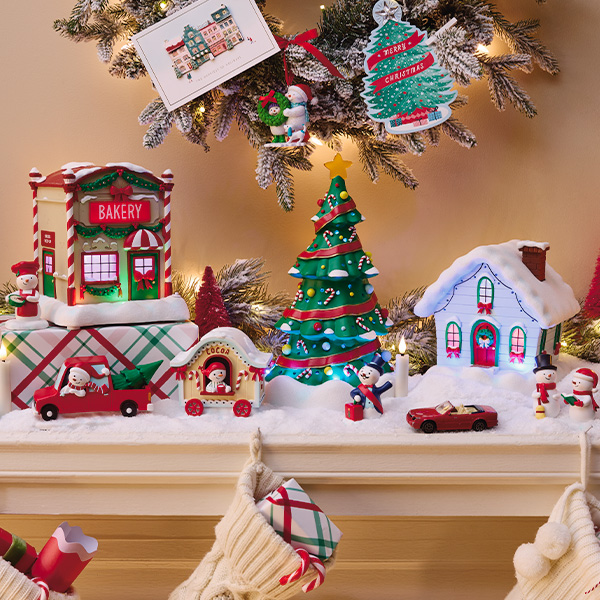 Festive holiday mantel display featuring miniature Christmas village decorations, wrapped gifts with plaid patterns and red ribbon, a decorated Christmas tree, toy cars, and snowy houses. Above the mantel, two holiday card ornaments hang from a flocked tree branch, including a Christmas tree-shaped design with the word “Christmas” and a card illustrated with a festive storefront. Knit stockings filled with gifts hang below, completing the cozy holiday scene.